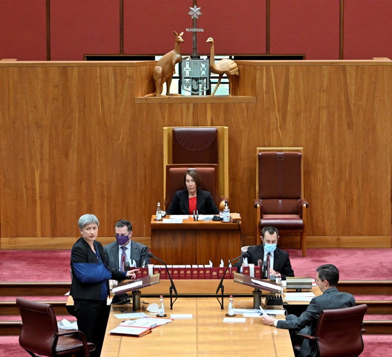 The President of the Senate sits in a large red chair. In front of her, the Clerks sit at a table with books on it. 
