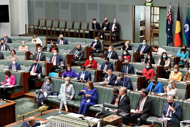 People in suits sit on green benches. One is standing and speaking.