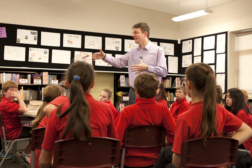 A teacher and students in a classroom. The students are sitting down and wearing red uniforms. The teacher is standing.